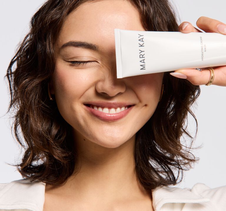 Woman smiling and holding a tube of Mary Kay&reg; cream in front of one side of her face, promoting the Skincare 101 section of the website.